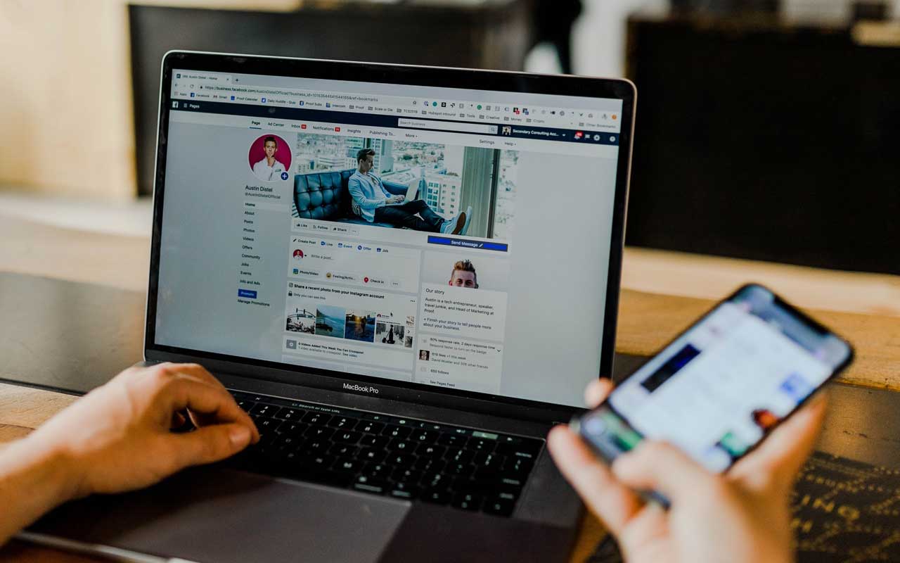A person holds a smartphone and scrolls through Facebook on their laptop.