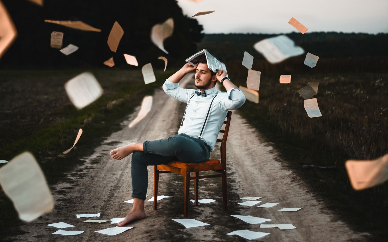 A man sits on a wooden chair in the middle of a dirt road. He covers his head with a book, protecting himself from a large number of falling papers.