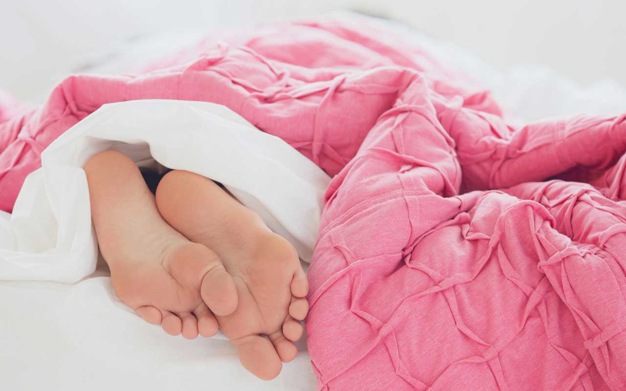 A pair of feet stick out from under a pink comforter on a bed. Getting enough sleep is key to maintaining your memory.