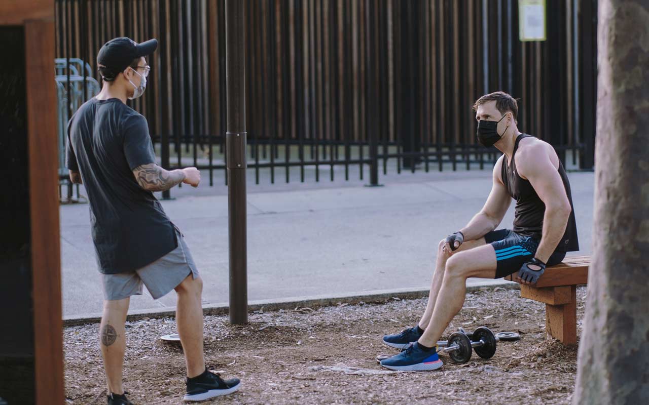 Two men wearing masks stand outside socializing while one man works out.
