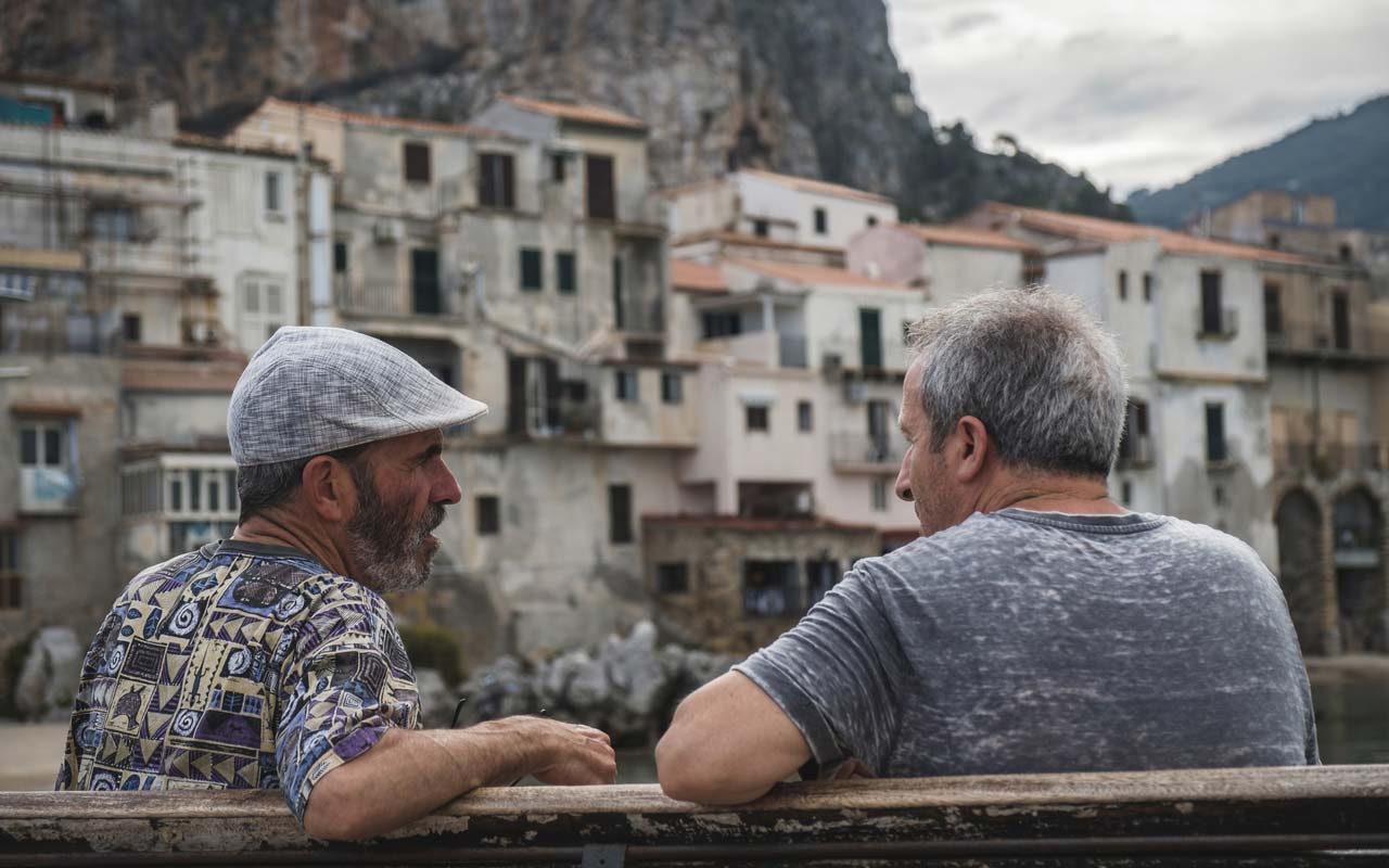 Two men sit on a bench speaking. 