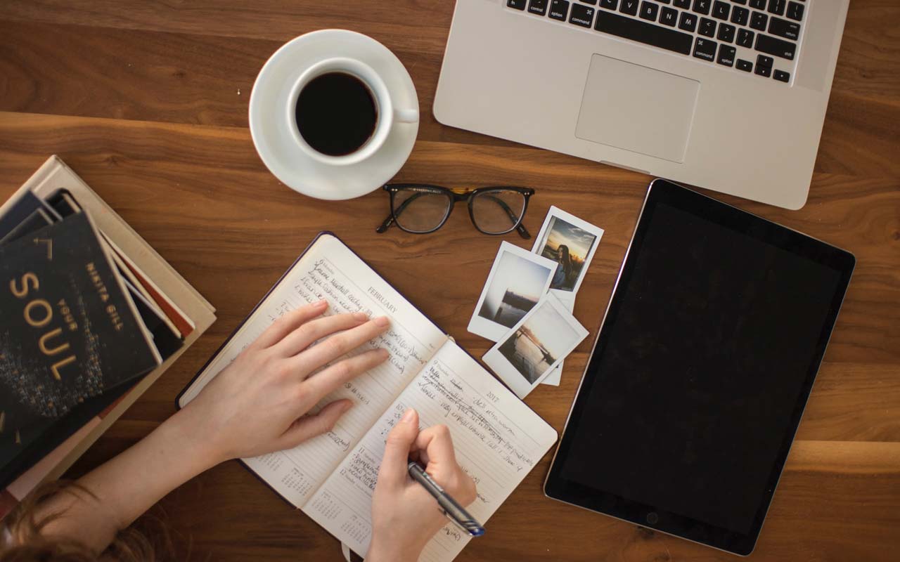 A person writes in a notebook, on a table with a laptop, coffee cup, and books.