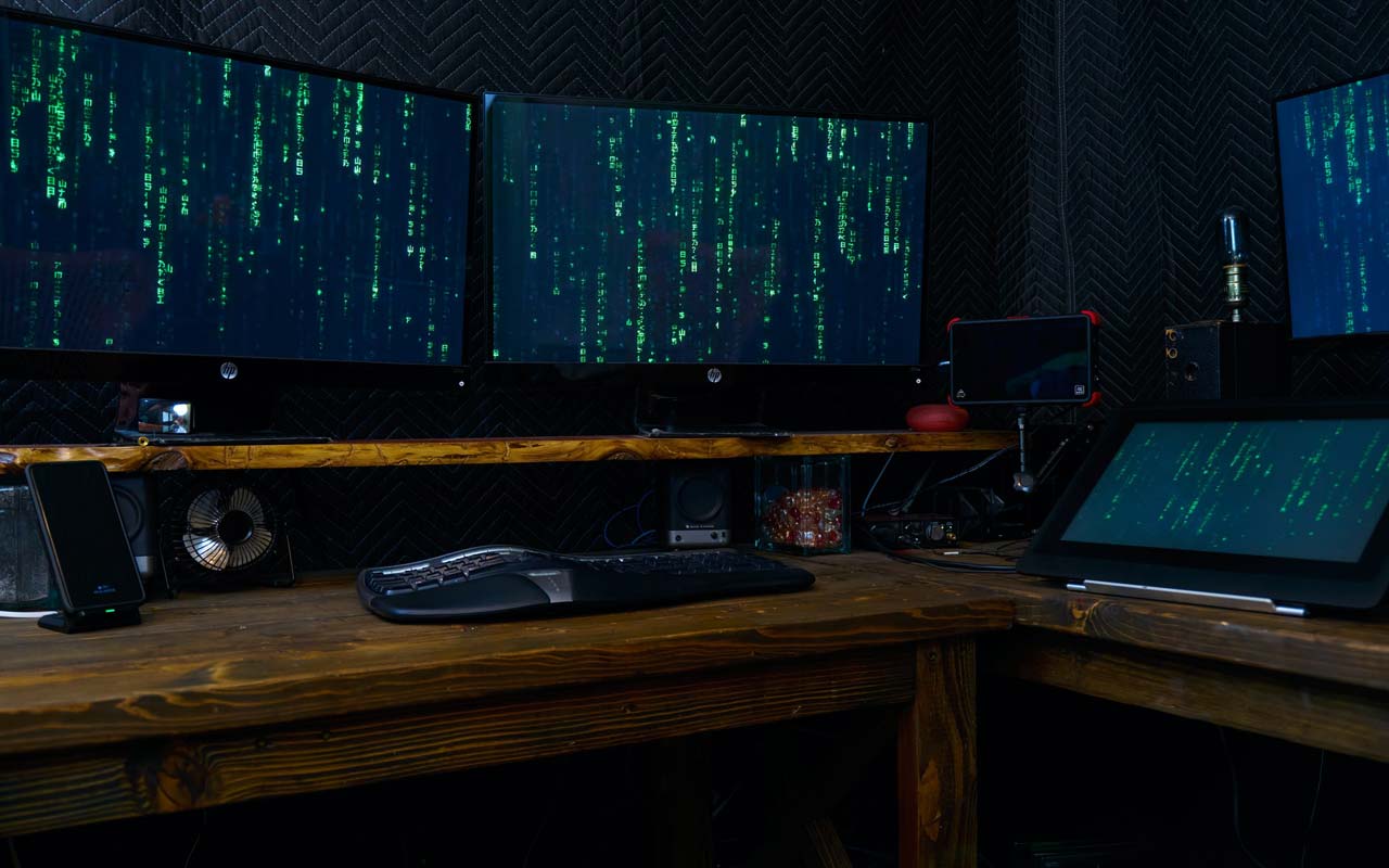 Multiple computer screens sit on a desk, running code that looks like the type used in the movie The Matrix.
