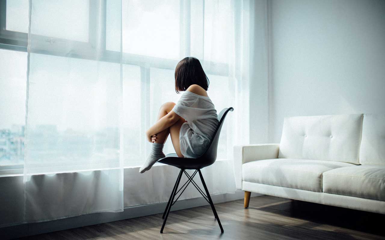 A woman sits on a chair with her knees drawn up to her chest. She looks out through sheer curtains through large glass windows.